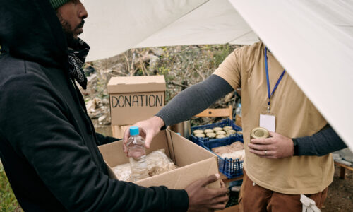 Young refugee man holding box while volunteer putting there food and water for the family of migrants